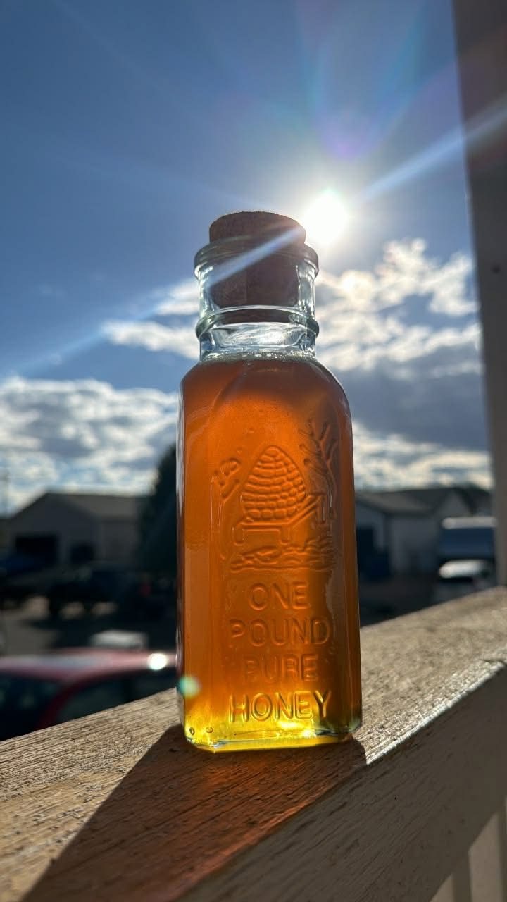 A one-pound embossed glass honey bottle with a cork stopper, glowing amber in the sun on a porch railing